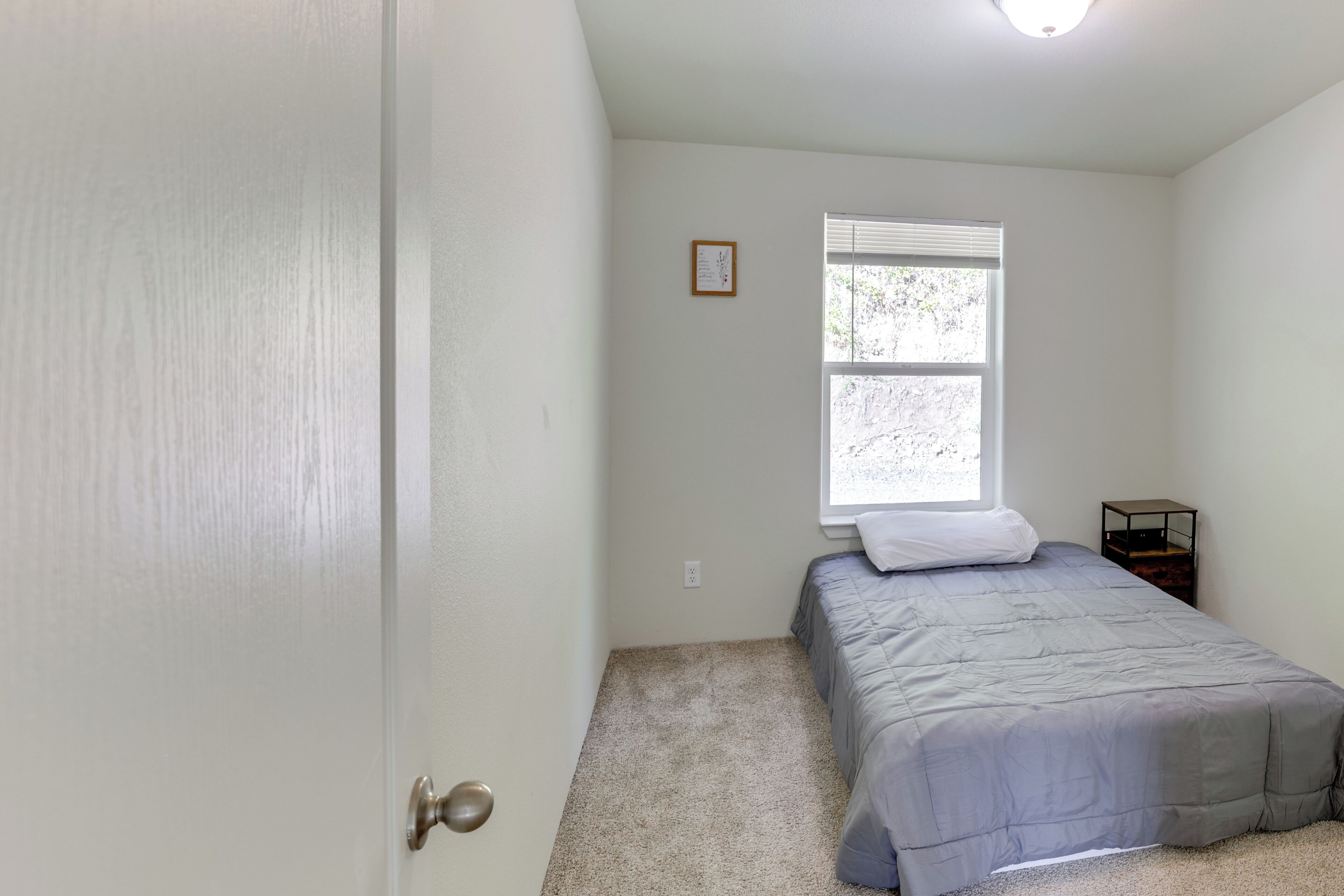 A bedroom with a single bed, a window with blinds, a small bedside table, and a door.