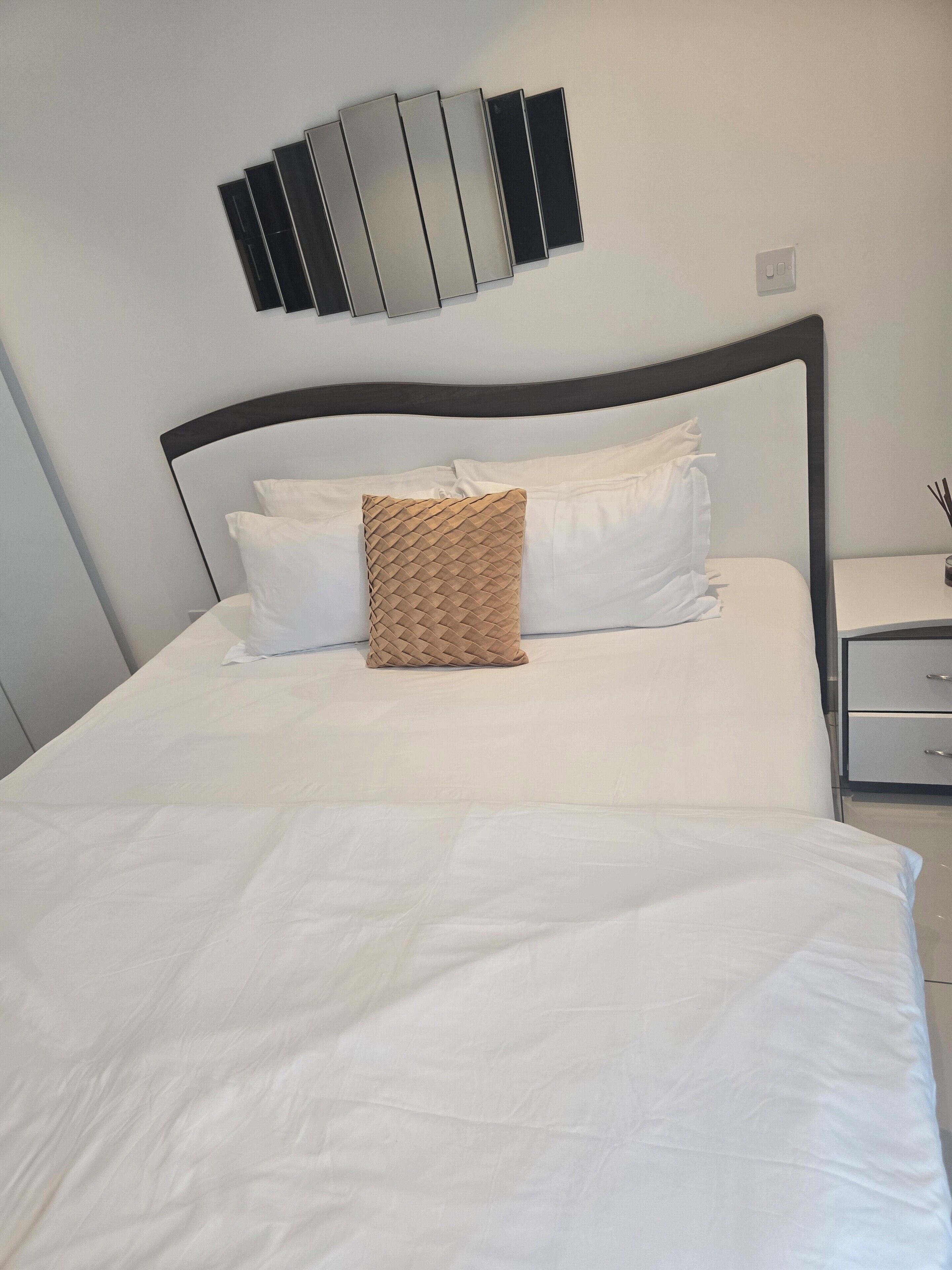 Una cama bien tendida con sábanas blancas y un cojín marrón texturizado, una lámpara moderna empotrada en la pared y una mesita de noche con cajón.