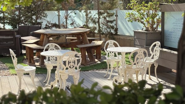 An outdoor patio with white metal tables and chairs, surrounded by wooden furniture and greenery. - Weibdeh Heights (Amman)