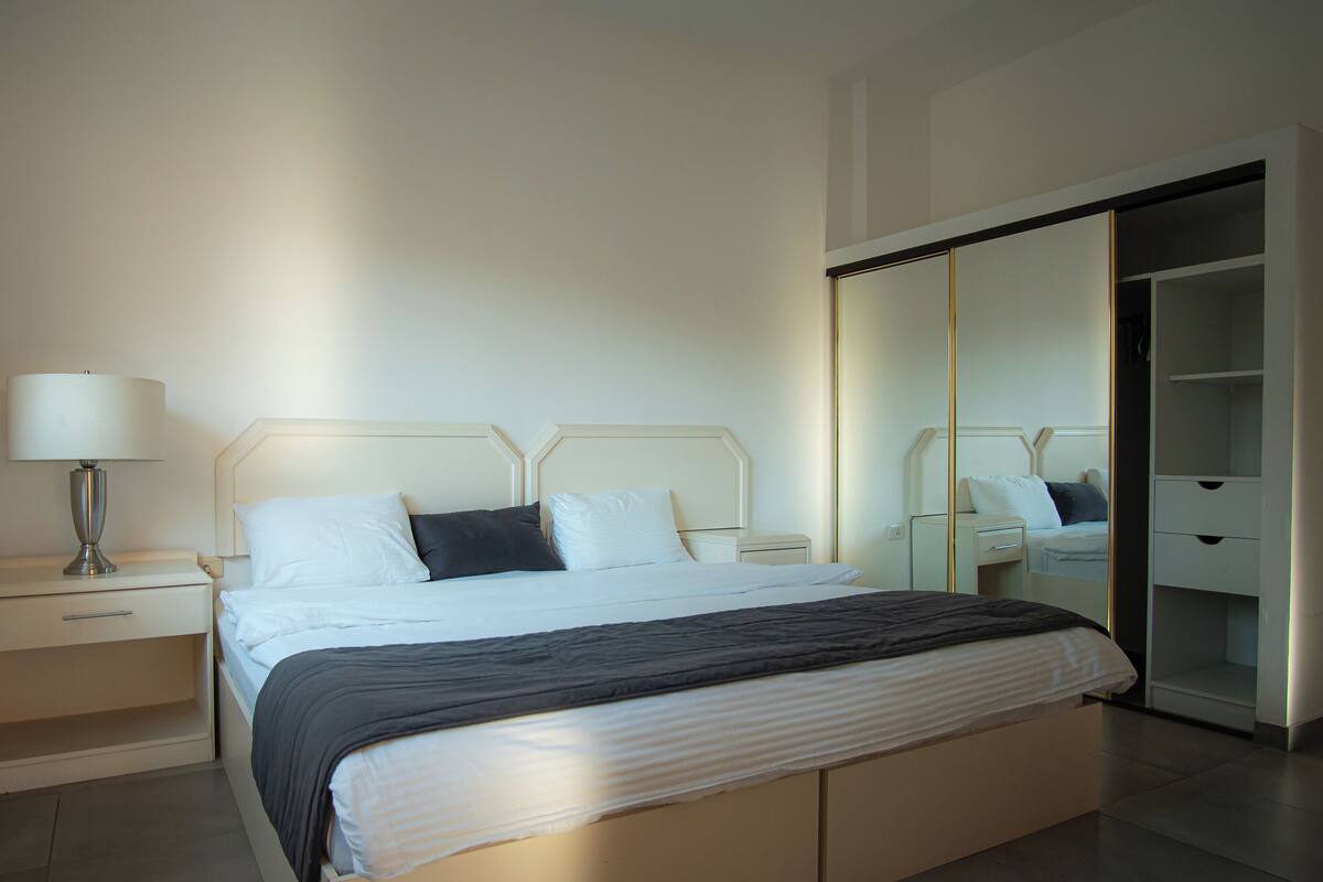 A modern hotel room with a neatly made bed, bedside tables, a lamp, and a large mirror.