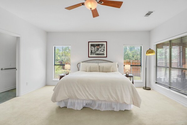A bedroom with a bed, a ceiling fan, a framed picture, and large windows with blinds. - Charming Kyle Retreat with Pool, Porch & Walk to Downtown (Kyle)