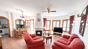 Una sala de estar con chimenea, televisor y comedor, con una cocina al fondo.