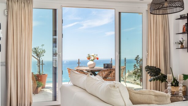 Uma sala de estar com um sofá, uma mesa com um vaso de flores e vista para o oceano através de grandes portas de vidro.