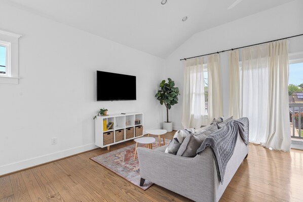 A modern living room with a grey sofa, a flat-screen TV, a small white table, and a large window with sheer curtains. - Solace One | Houston (Houston)