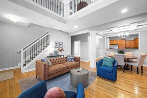 Un salon spacieux avec un canapé marron, des fauteuils bleus et une table basse ronde. Un escalier avec une rampe mène à un niveau supérieur.