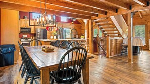 Un vaste espace à manger en bois, avec une grande table et des chaises, une cuisine à l’arrière-plan et un escalier menant à un niveau supérieur.