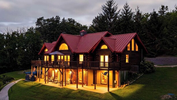 Une grande cabane en rondins dotée de nombreux balcons et d’une véranda qui fait le tour de la maison, entourée d’une pelouse bien entretenue et d’arbres denses.