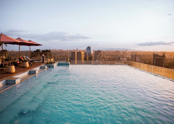 A rooftop infinity pool with a cityscape view, lounge chairs, and umbrellas.