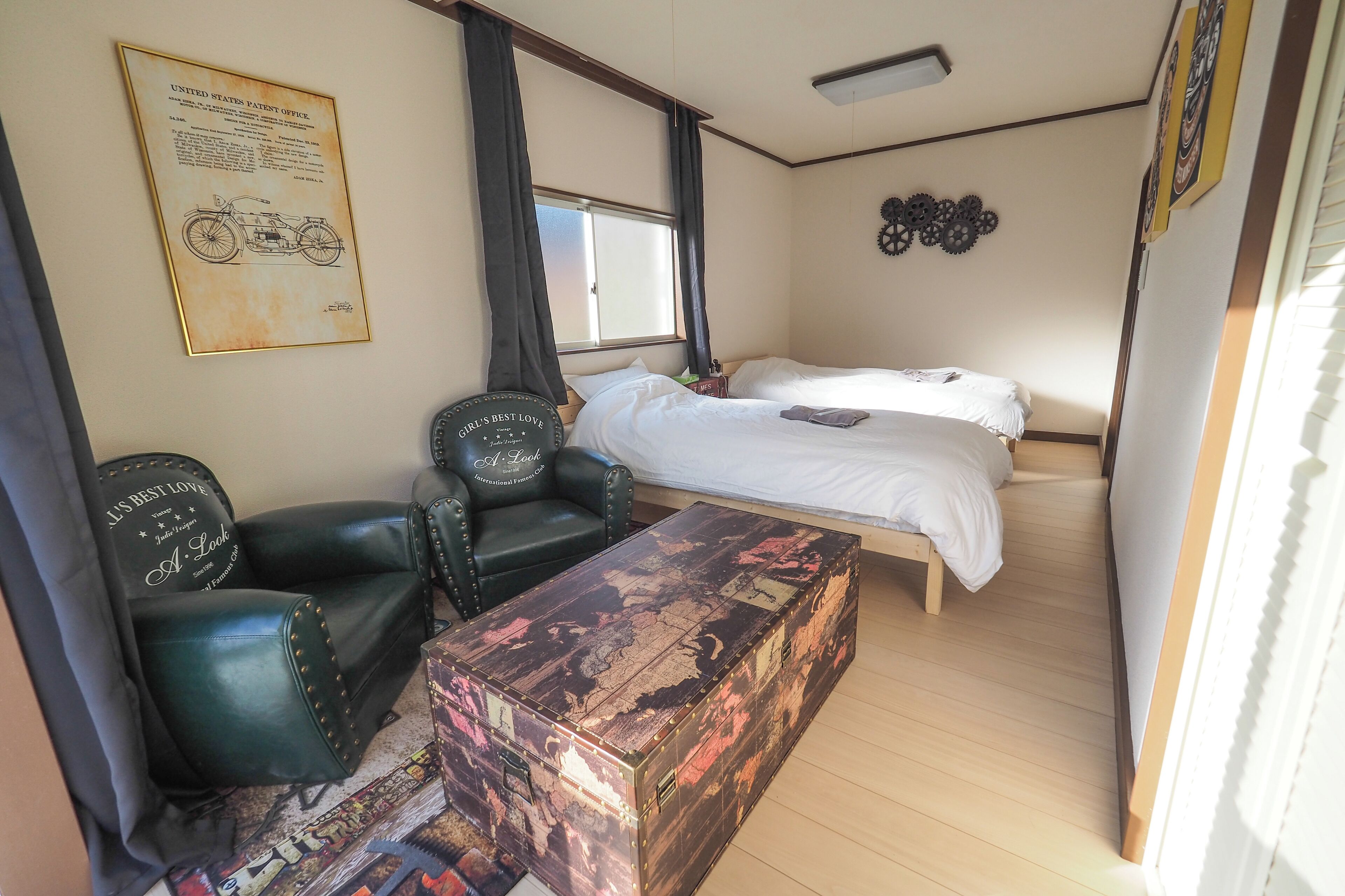 Una habitación con una cama, un escritorio, dos sillones de cuero, un baúl antiguo y un plano de motocicleta colgado en la pared.