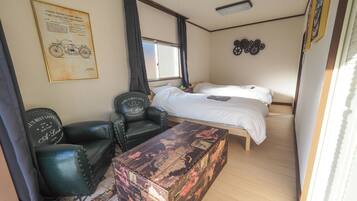 Una habitación con una cama, un escritorio, dos sillones de cuero, un baúl antiguo y un plano de motocicleta colgado en la pared.
