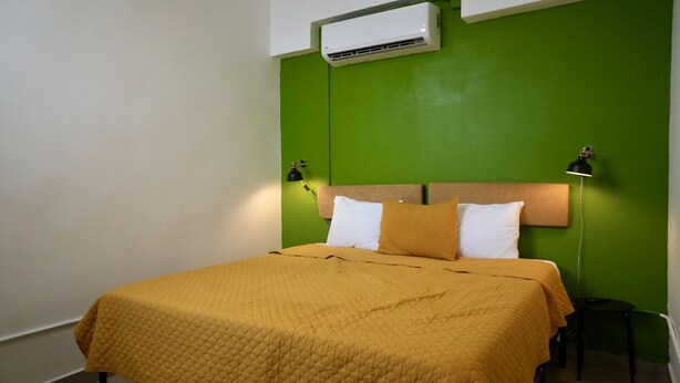 A hotel room with a bed, green walls, a white air conditioner, and a nightstand with a lamp.