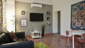 A living room with a black leather sofa, a white side table, a flat-screen TV mounted on the wall, a dining table with red chairs, and a large abstract painting on the wall.