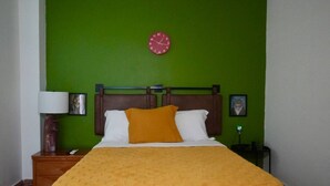 Ein Hotelzimmer mit grüner Wand, einem hölzernen Kopfteil, einem Bett mit orangefarbener Decke, einer roten Uhr und einem Nachttisch mit Lampe und Bilderrahmen.