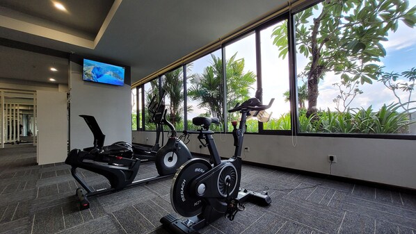A gym with stationary bikes and a treadmill, large windows with a view of greenery, and a television on the wall. - Palam Mansion Citra Plaza Nagoya (Batam)