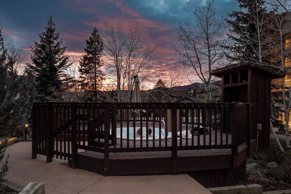 Un espace avec un bain à remous, doté d’une terrasse en bois et d’une petite structure en bois, le tout entouré d’arbres, avec un coucher de soleil en arrière-plan.