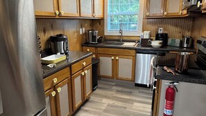 A compact kitchen with wooden cabinets, stainless steel appliances, a window with blinds, and a fire extinguisher.