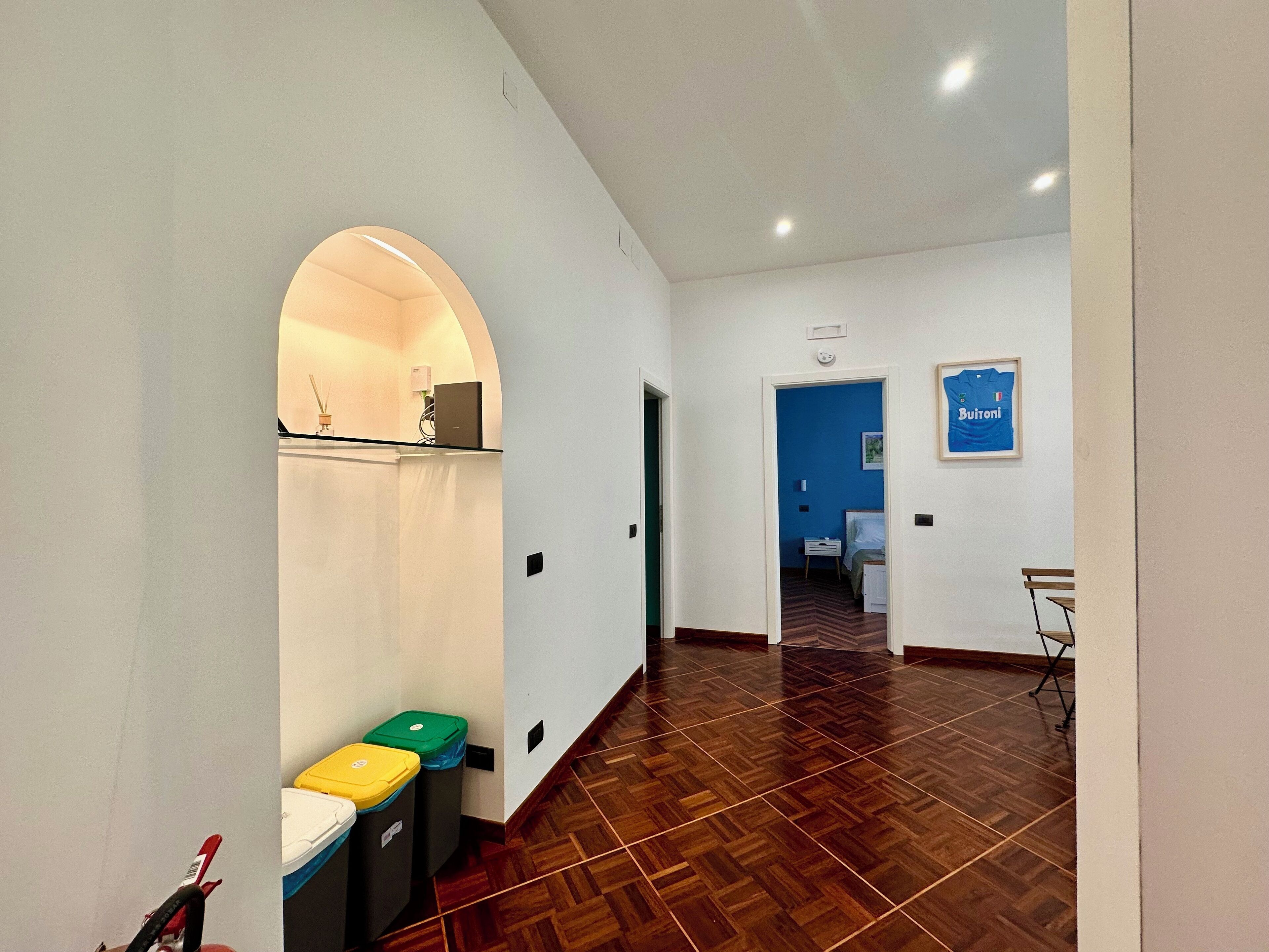 A hotel corridor with a wooden floor, a blue wall, and a framed picture. There are trash bins and a small table with a laptop on it.
