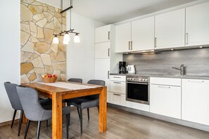 Une cuisine moderne avec des meubles blancs, un mur d’accent en pierre, une table à manger en bois et des chaises recouvertes d’un tissu gris.