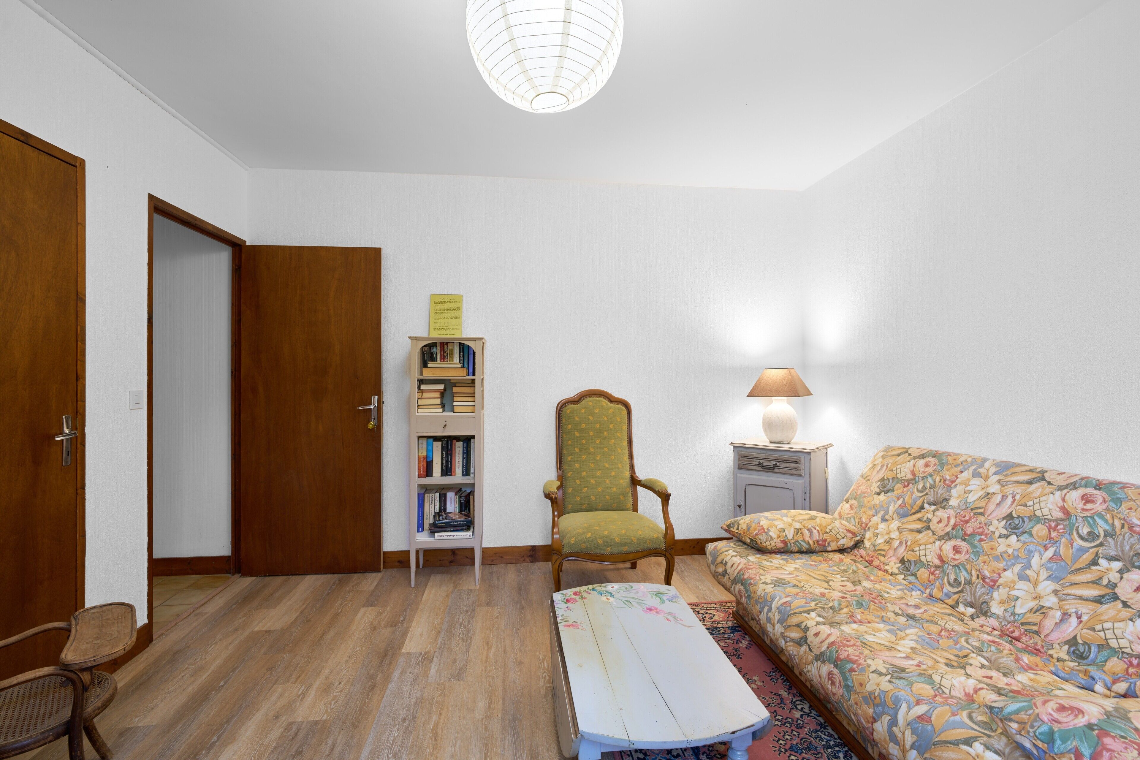 Una habitación con suelo de madera, un sillón verde, una estantería, una cama con colcha estampada y una lámpara en la mesita de noche.