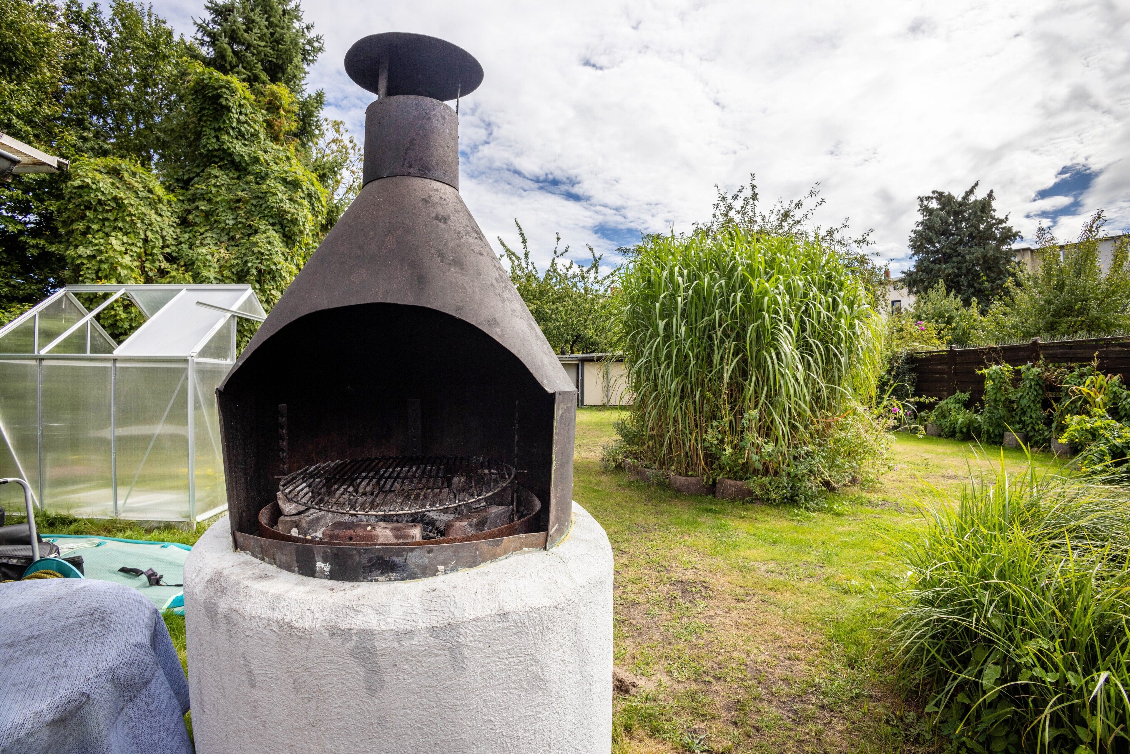 A concrete outdoor grill with a chimney, surrounded by greenery and a greenhouse in the background.