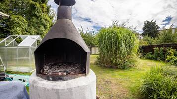 A concrete outdoor grill with a chimney, surrounded by greenery and a greenhouse in the background.
