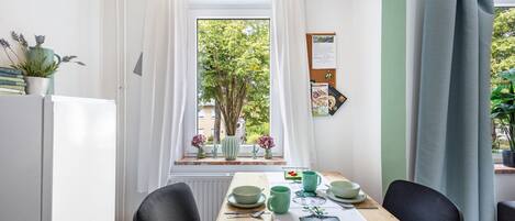 Un comedor con una mesa dispuesta para dos personas, una ventana con cortinas, un tablón de anuncios y una planta en maceta en el alféizar.