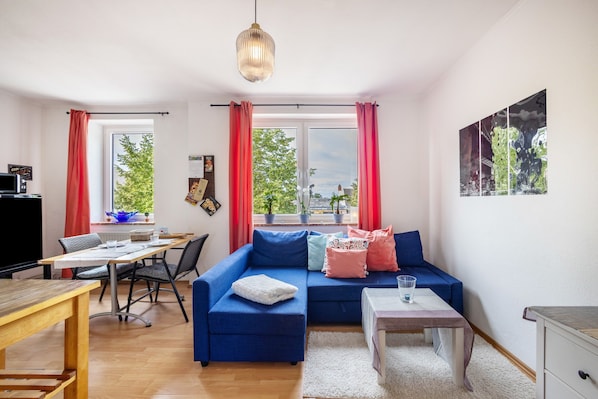 Ein Wohnzimmer mit einem blauen Sofa, einem Holzesstisch, einem Fernseher und einem Fenster mit roten Vorhängen.