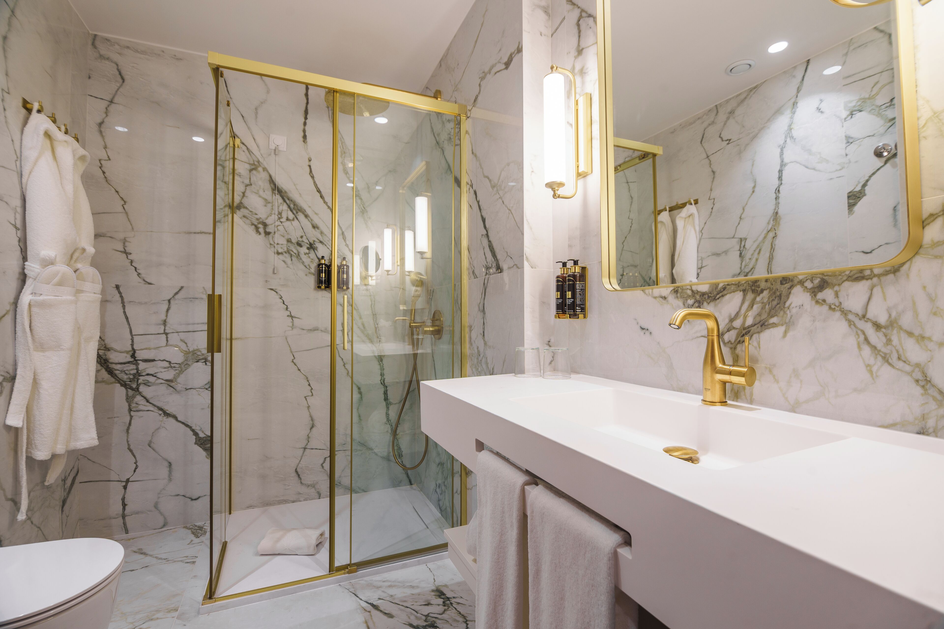 Un baño moderno con ducha envuelta en vidrio, tocador blanco con grifo dorado y paredes de mármol.