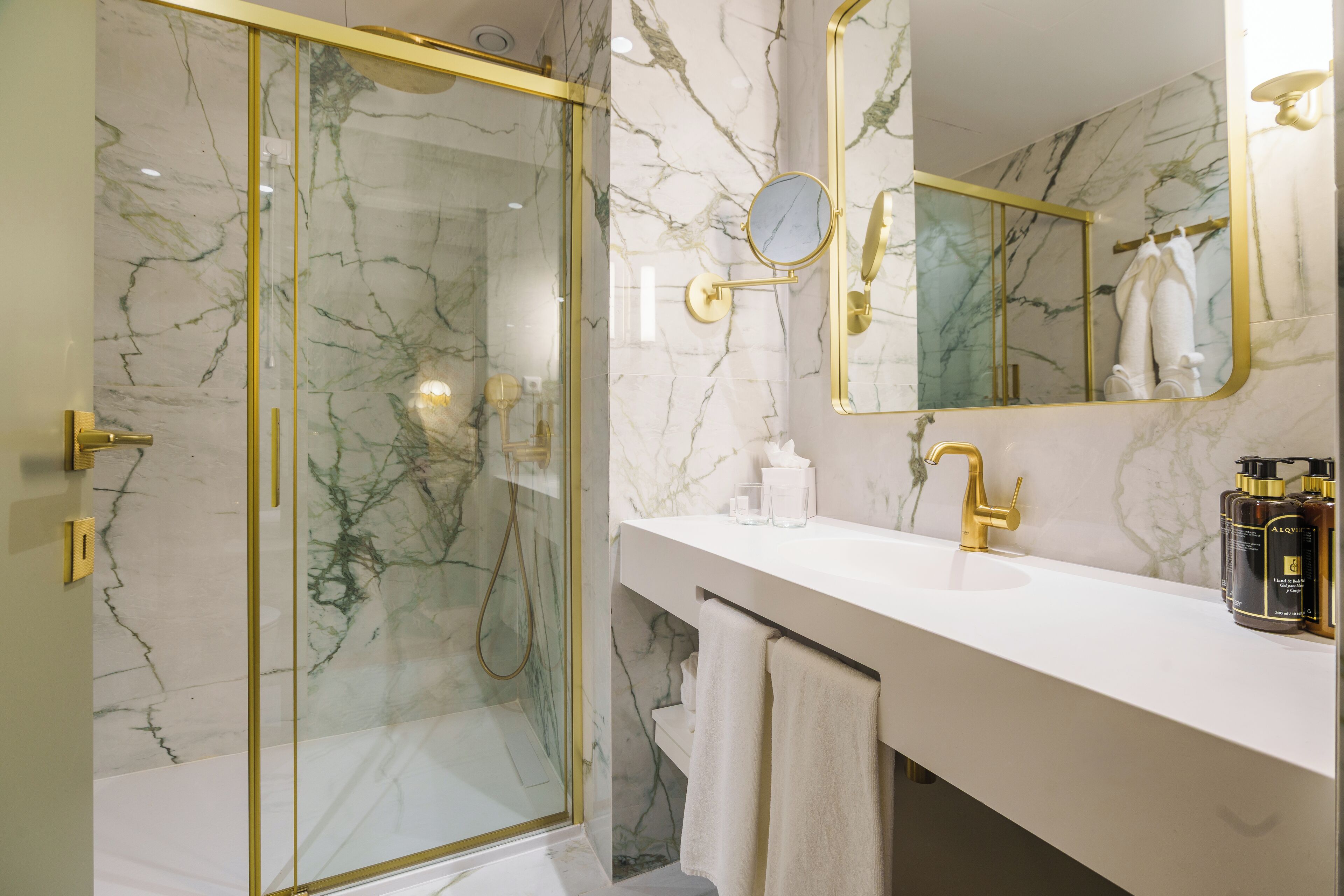 Un baño moderno con ducha envuelta en vidrio, salpicadero de mármol, grifo dorado y tocador con artículos de aseo.