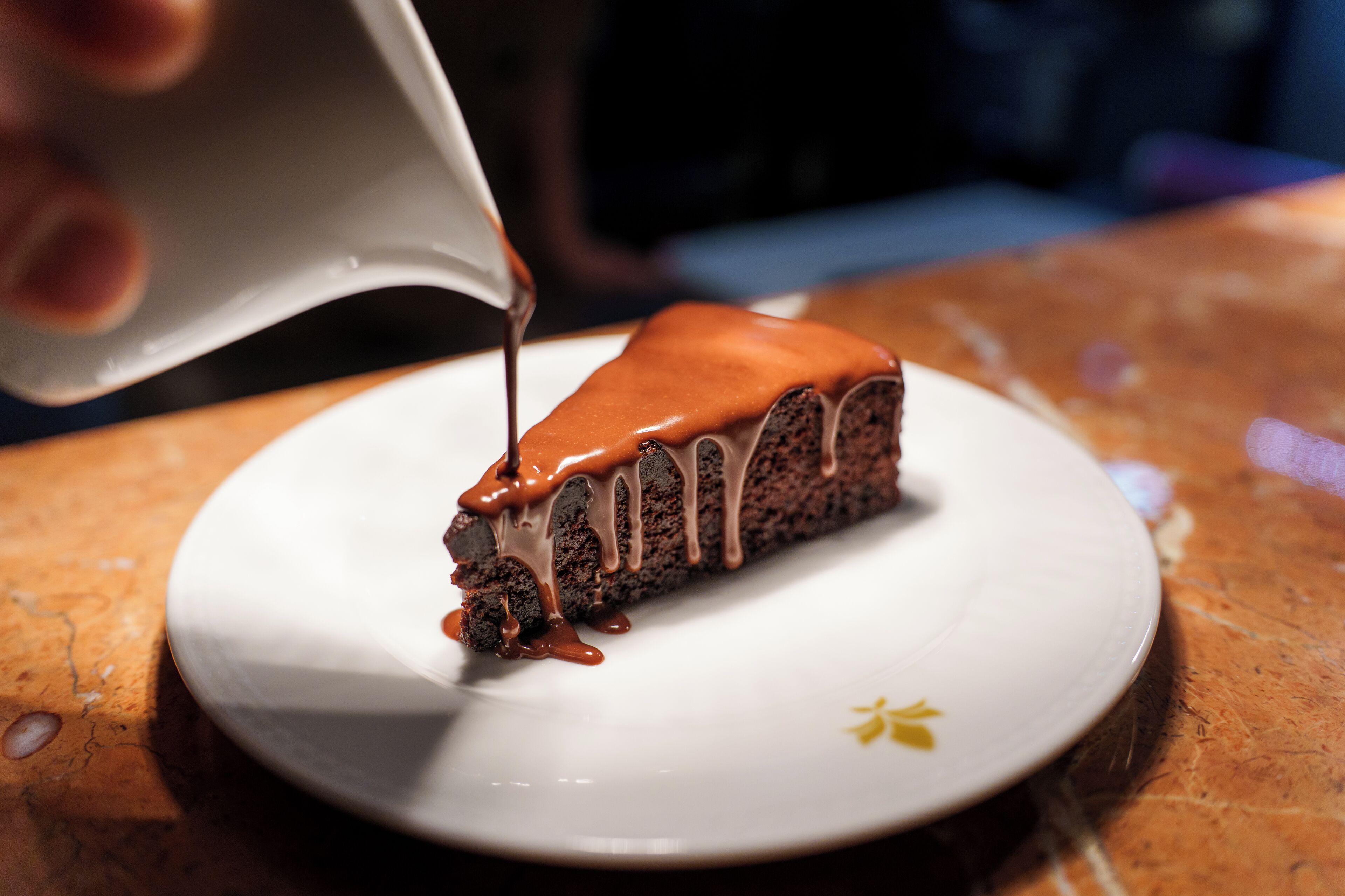 A slice of chocolate cake with chocolate sauce being poured over it.