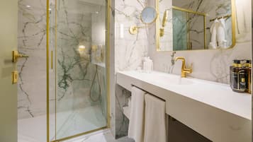 A modern bathroom with a glass-enclosed shower, a marble backsplash, a white vanity with gold fixtures, and a large mirror.