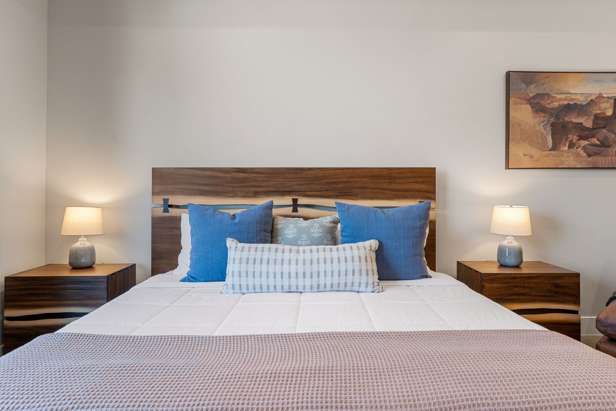 A neatly made bed with a wooden headboard, two bedside tables with lamps, and a painting on the wall.
