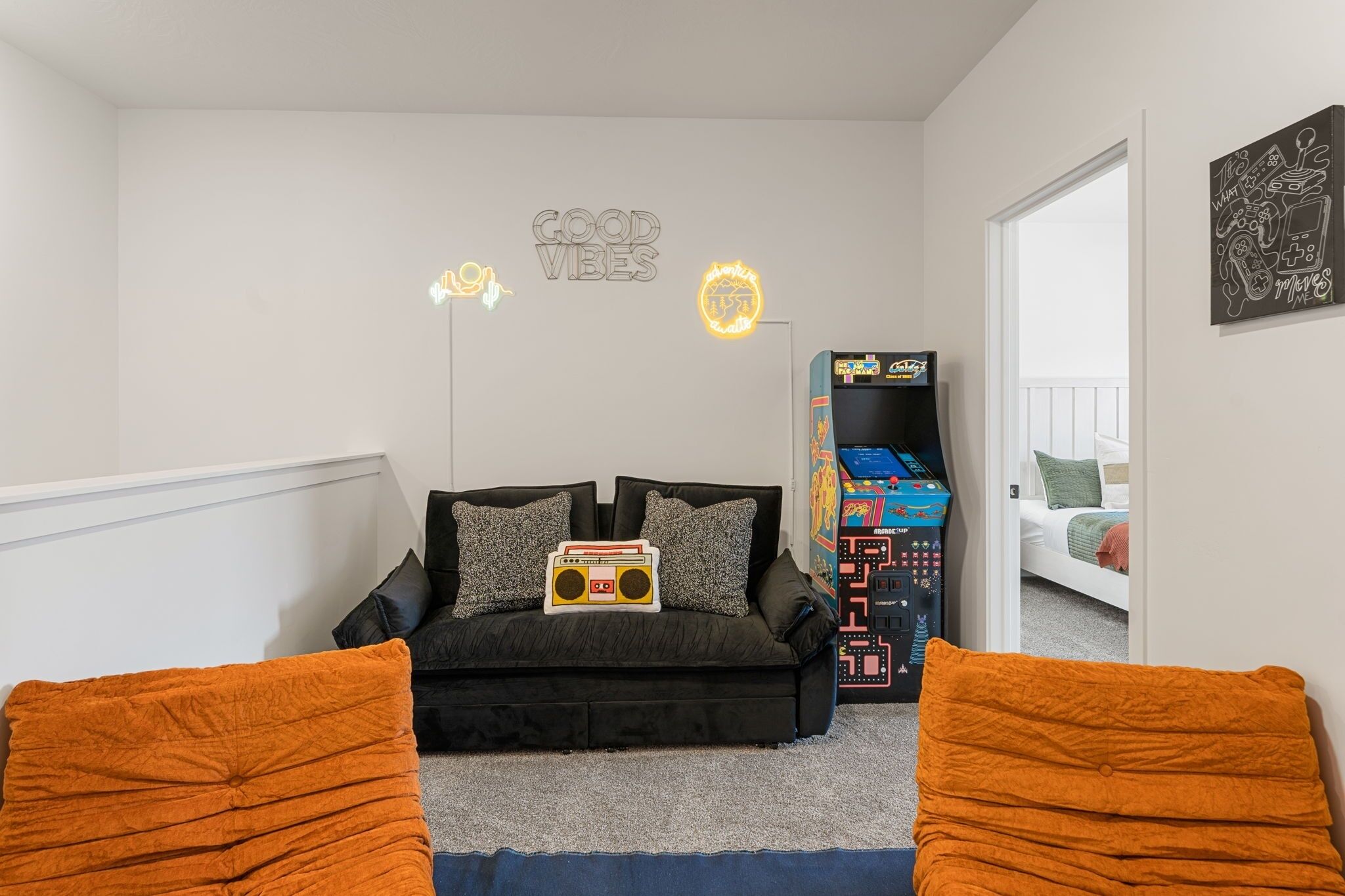 Un dormitorio con sofá negro, cojines naranjas, un letrero de neón y un juego de mesa.
