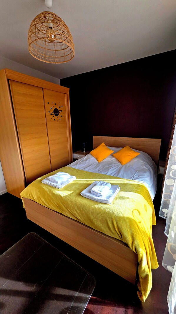 Una cama bien hecha con un edredón amarillo y dos almohadas naranjas, un armario de madera y una lámpara colgante.