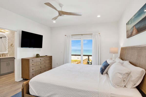 A bedroom with a bed, a ceiling fan, a TV, a window with a view of the beach, and a wooden dresser.