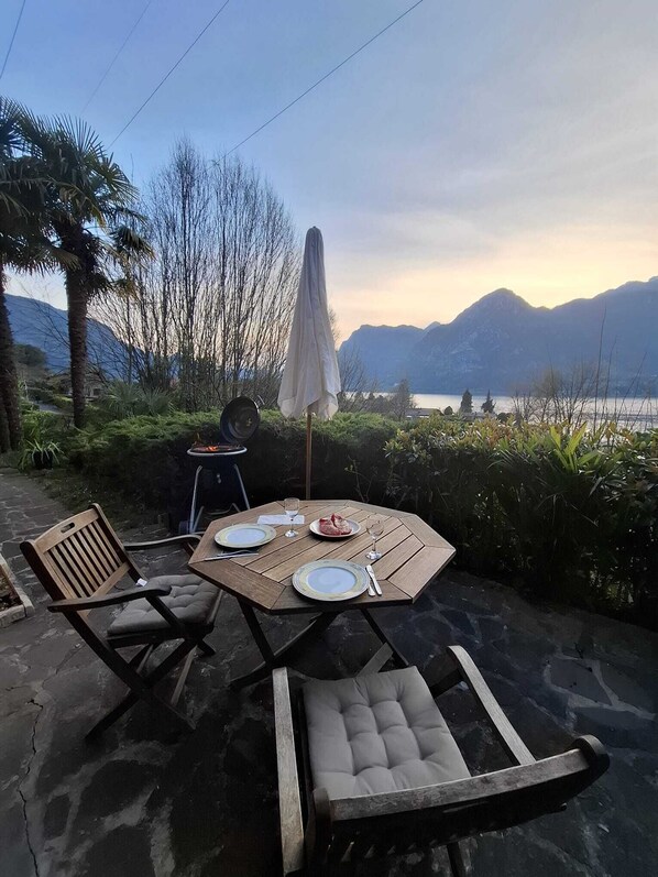 An outdoor dining area with a table set for two, chairs, a grill, and a view of mountains and a lake. - Casa Salvia: Lakeside Retreat with Stunning Mountain Views (Vesta)