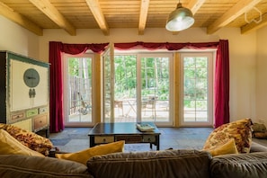 Ein gemütliches Zimmer mit Holzbalkendecke, einem Vintage-Schrank, einer Couch und einem Tisch. Große Fenster mit roten Vorhängen bieten einen Blick ins Freie.