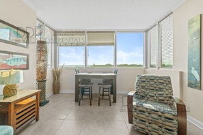 A dining area with a table and chairs, a patterned armchair, and decorative items. - Ocean Club 403 | Enjoy Views of the Gulf! (Biloxi)