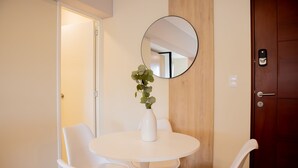 Una mesa pequeña y redonda, blanca, con cuatro sillas blancas, un florero blanco con follaje verde y un espejo redondo en la pared.