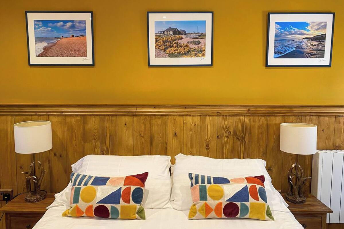 A hotel room with a wooden headboard, two bedside lamps, a bed with colorful geometric pillows, and three framed beach photographs on the wall.
