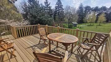 A wooden deck with outdoor furniture, including a round table and chairs, overlooking a grassy area with trees.