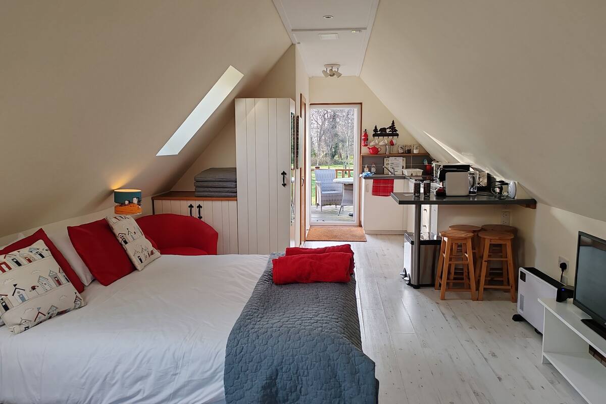 A bedroom with a bed, a red chair, a desk, and a kitchenette.