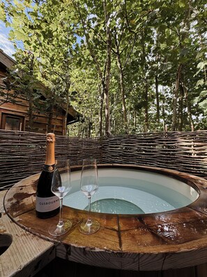 A wooden hot tub with a bottle of Gösburgen wine and two glasses on a wooden table, surrounded by trees.