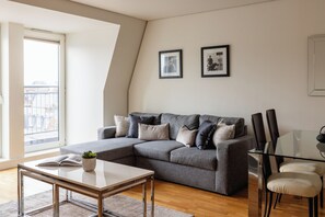 Un salon moderne avec un canapé d’angle gris, une table à manger en verre et une grande fenêtre donnant sur des immeubles.