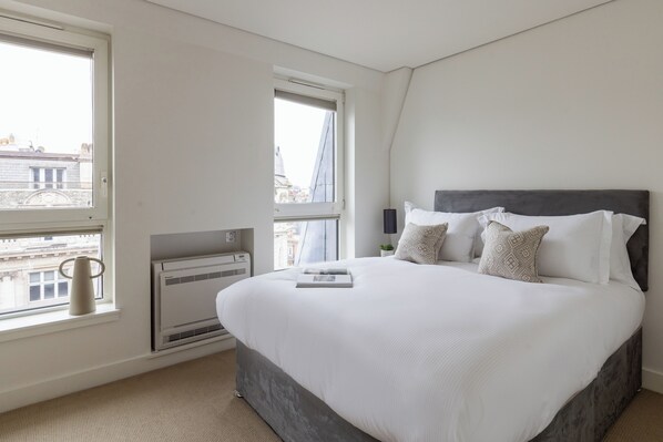 Een slaapkamer met een groot bed, een raam met uitzicht op gebouwen, een airconditioning en een nachtkastje met een lamp.