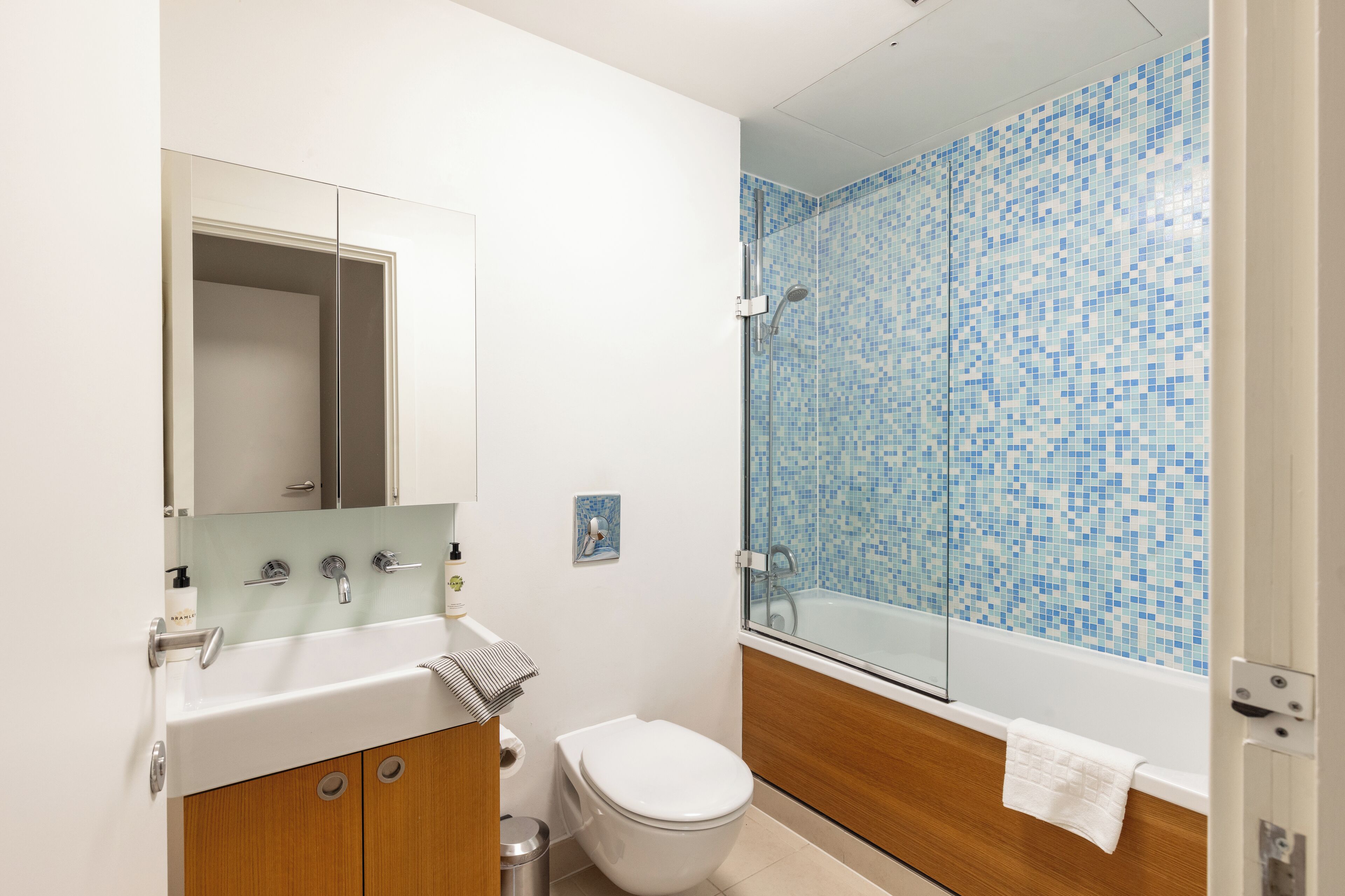 A bathroom with a white sink, mirror, and toilet. It features a shower with blue mosaic tiles and a wooden vanity.