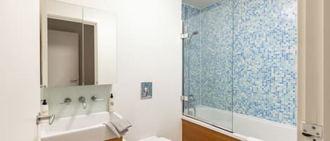 A bathroom with a white sink, mirror, and toilet. It features a shower with blue mosaic tiles and a wooden vanity.