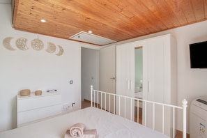 A bedroom with a wooden ceiling, a bed with a white headboard, a white dresser, a wardrobe, and a flat-screen TV mounted on the wall.