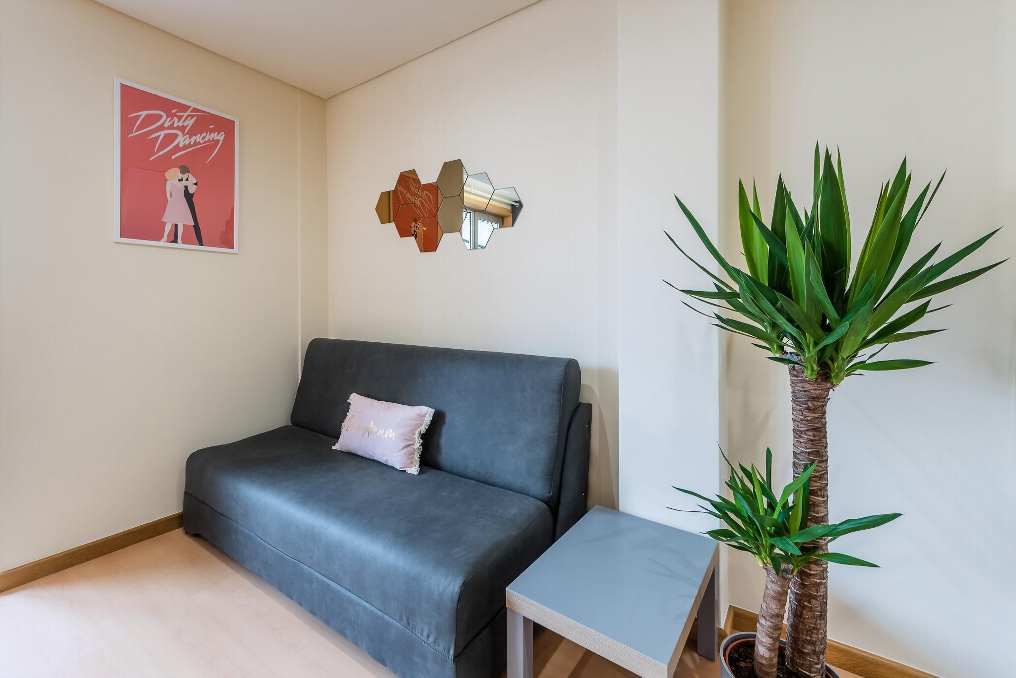 A room with a grey sofa, a small side table, a potted plant, a wall-mounted abstract art piece, and a poster on the wall.