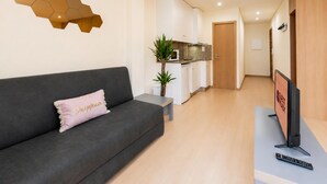 Uma sala de estar moderna com um sofá cinzento, uma mesa pequena, uma TV e uma cozinha ao fundo.